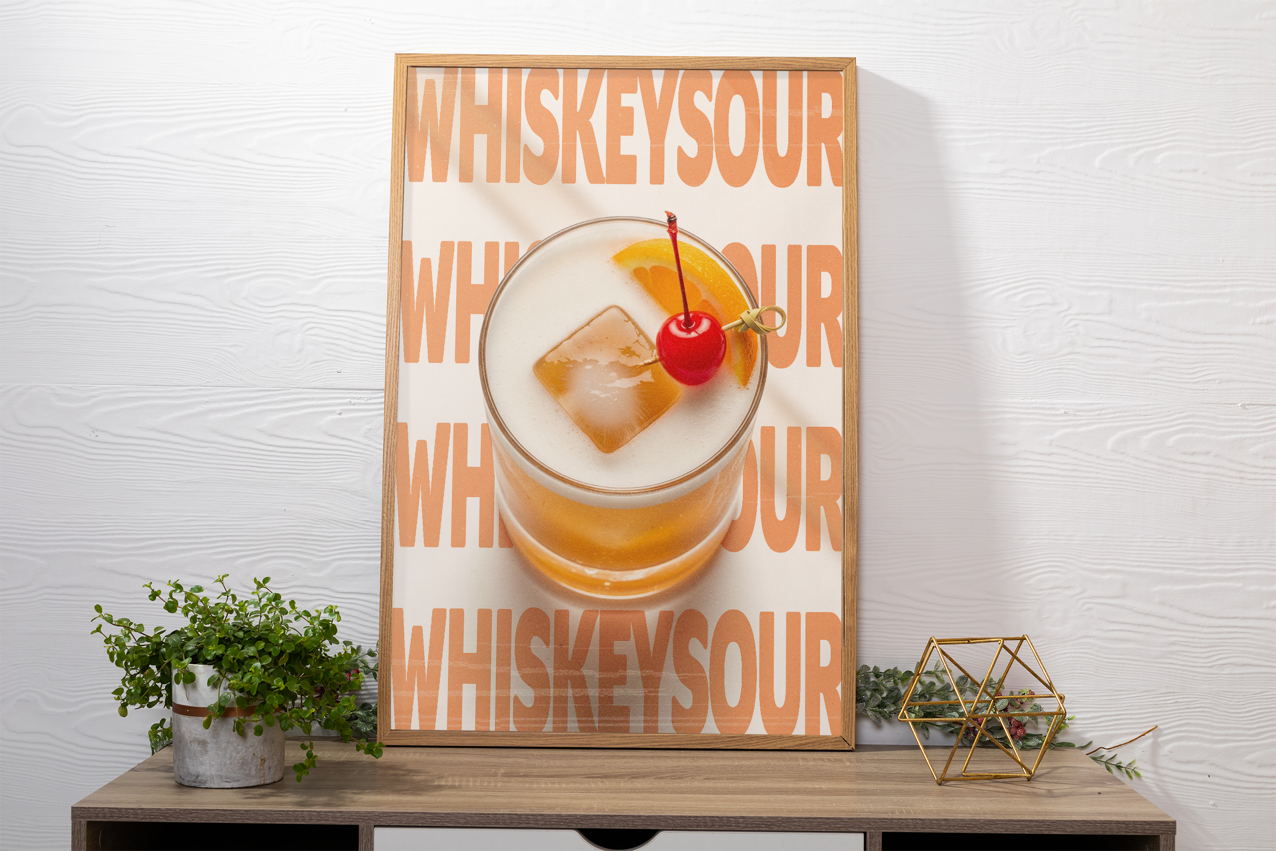 Framed poster of a Whiskey Sour cocktail garnished with a cherry and orange slice, with repeating 'WHISKEYSOUR' text, on a white textured background above a dark dresser.