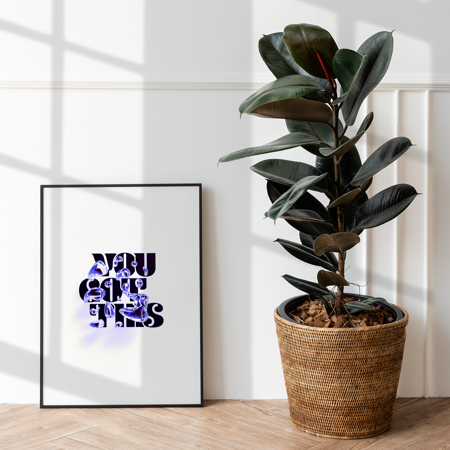 A black-framed poster with glowing blue and purple 3D text "YOU GOT THIS" resting on a light wood floor next to a large dark rubber plant in a woven basket.
